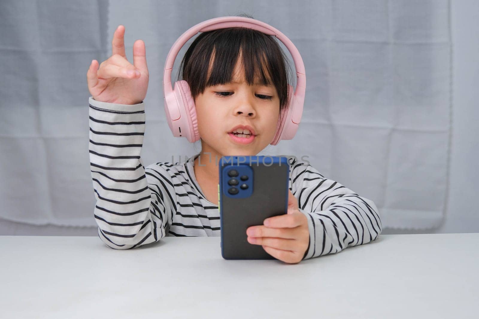 Cute elementary school girl wearing headphones holding a smartphone. Happy Asian girl studying online on smartphone or homeschooling, listening to music or playing games. by TEERASAK