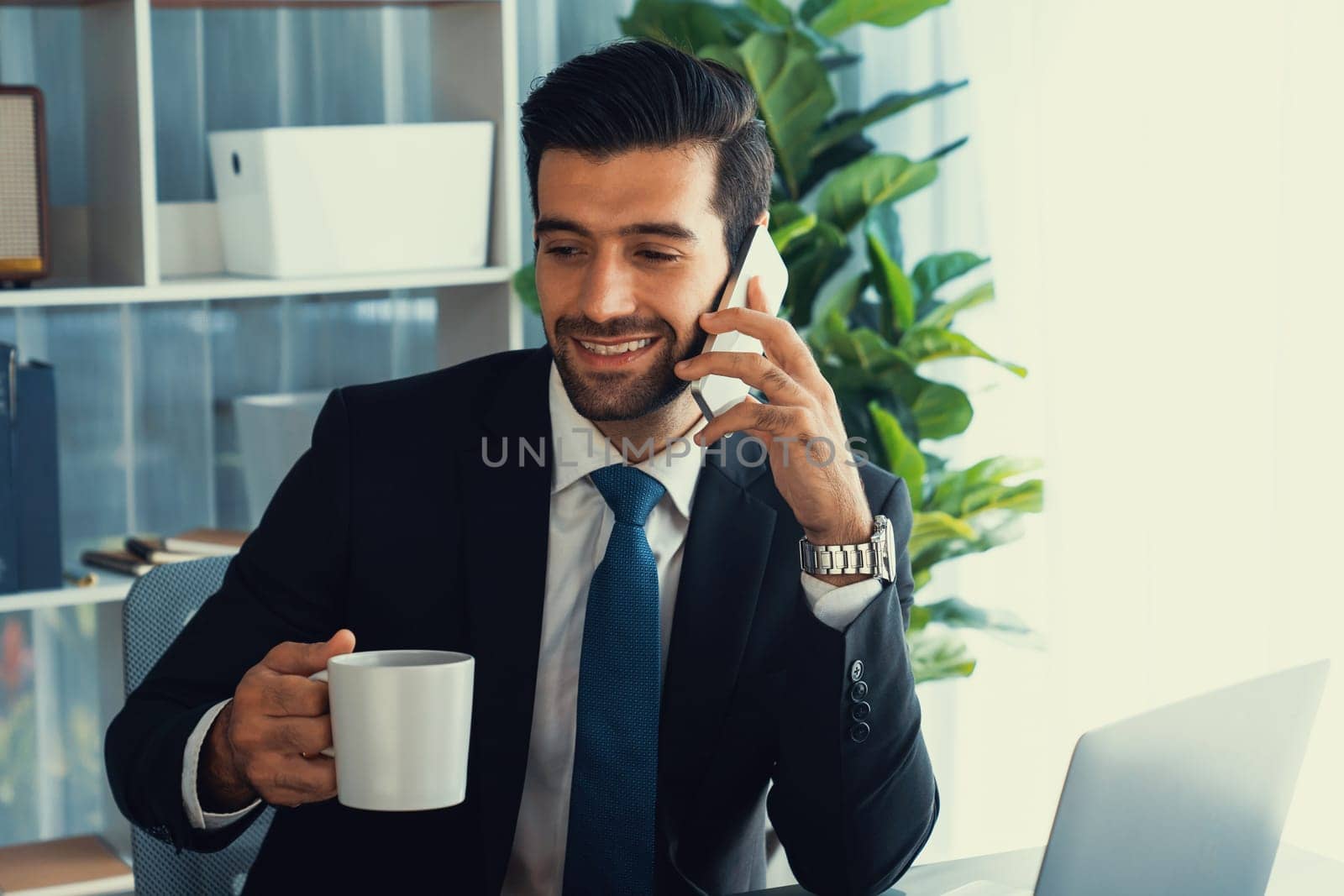 Businessman working in modern office workspace with cup of coffee in his hand while answering phone call making sales calls or managing employee. Fervent