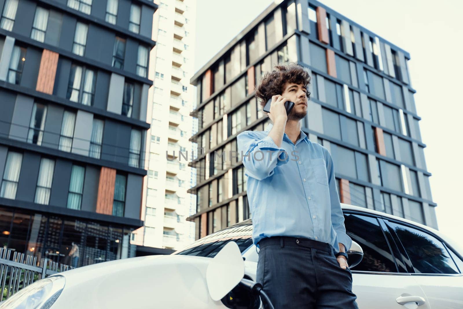 Progressive businessman talking on the phone, leaning on electric car recharging with public EV charging station, apartment condo residential building on the background as green city lifestyle.