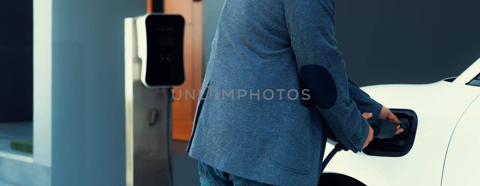 Progressive asian man install cable plug to his electric car with home charging station in the backyard. Concept use of electric vehicles in a progressive lifestyle contributes to clean environment.