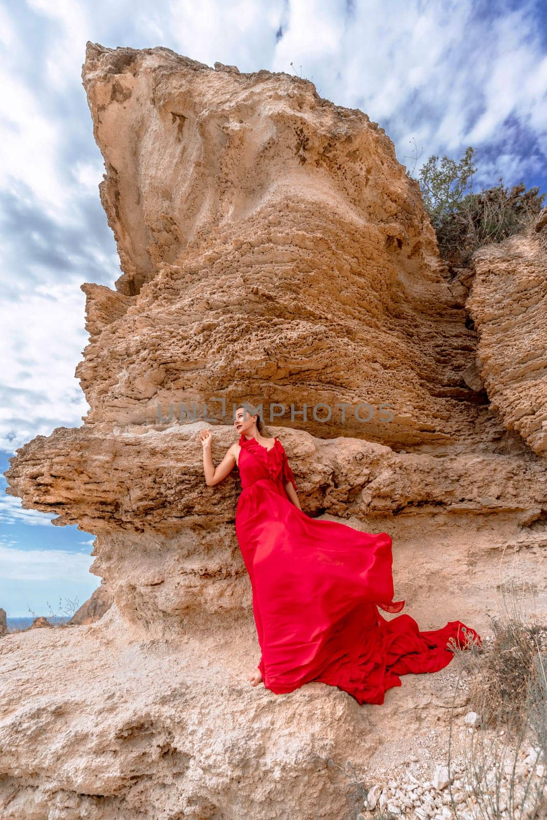 A woman in a red silk dress stands by the ocean, with mountains in the background, as her dress sways in the breeze