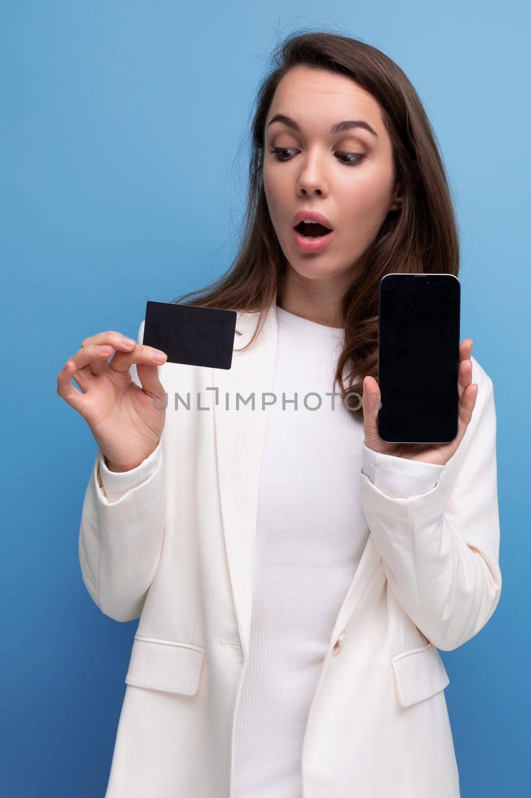surprised brunette young woman in a dress and jacket with a payment card for shopping and a smartphone.