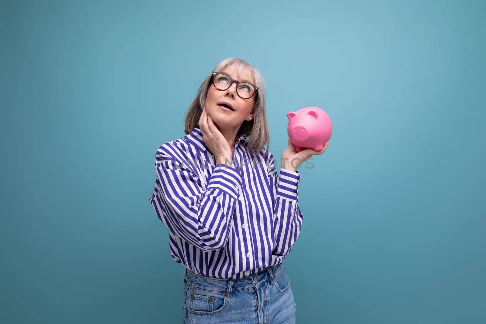 pensive 60s woman with gray hair holding piggy bank thinks about insurance on bright studio background by TRMK