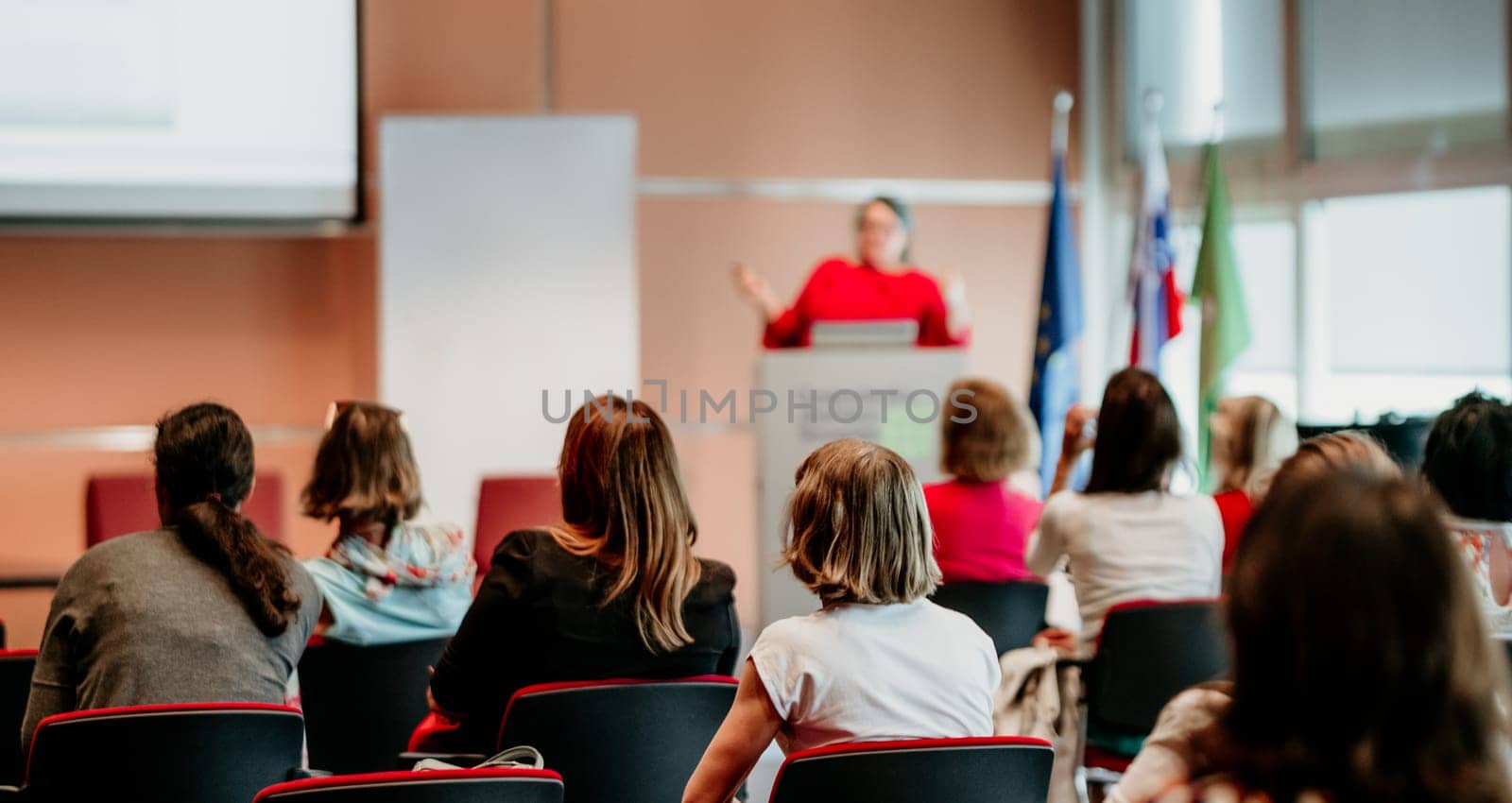 Business and entrepreneurship symposium. Female speaker giving a talk at business meeting. Audience in conference hall. Rear view of unrecognized participant in audience.