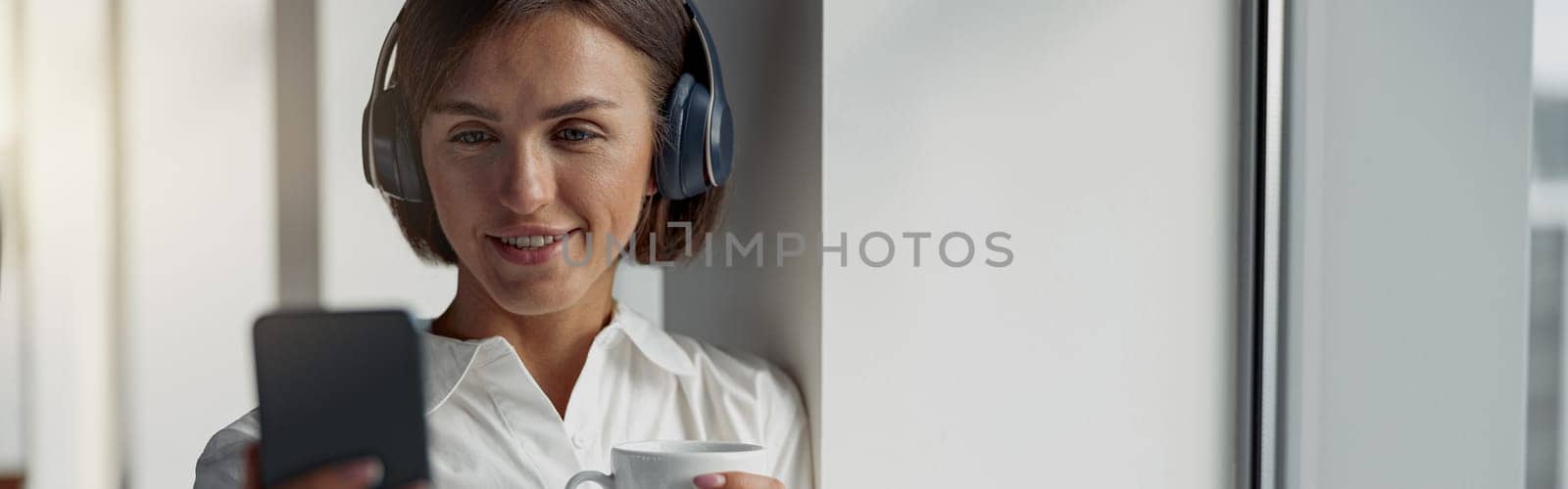 European business woman holding cup of coffee and using phone while standing near window at office