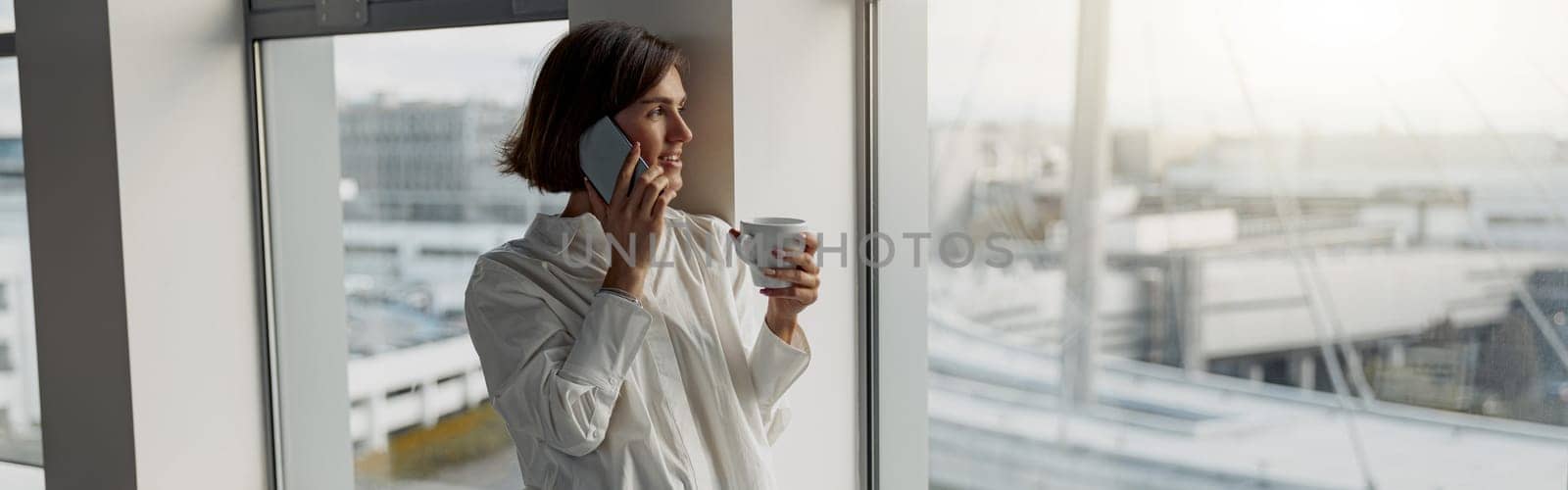 European business woman holding cup of coffee and talking phone while standing near window at office