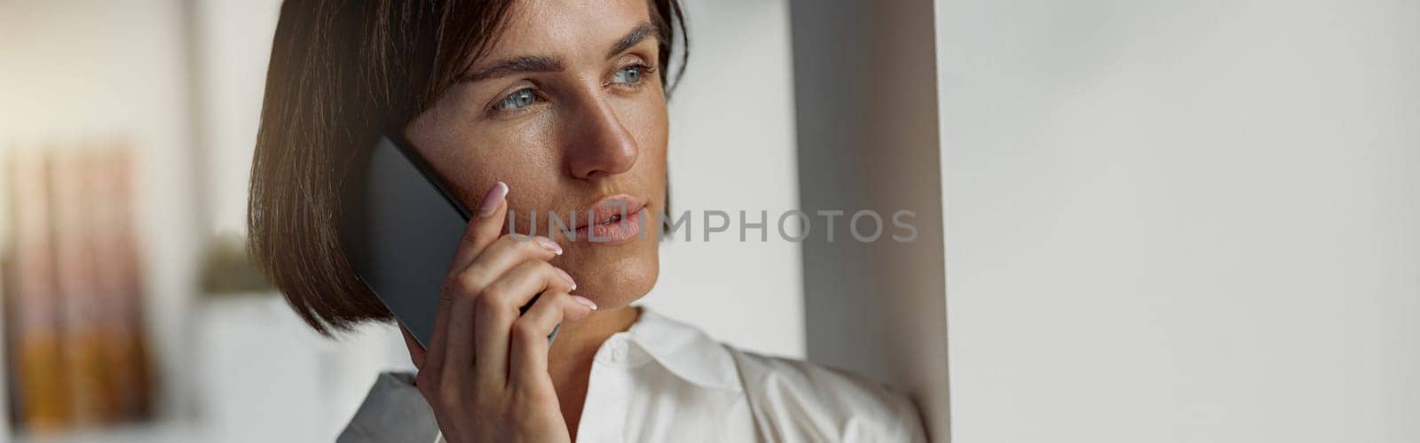 Business woman holding a cup of coffee and talking phone while standing near window at office by Yaroslav_astakhov