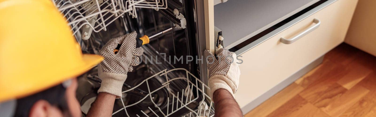 Close up of handyman in special clothing repairing dishwasher in modern kitchen by Yaroslav_astakhov