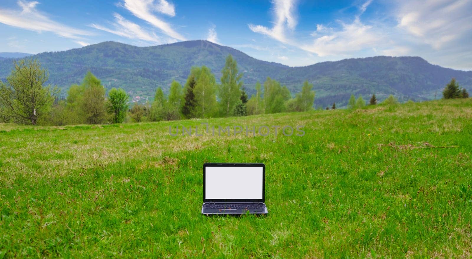 Green field and laptop lit by the mountains and blue sky. Open fresh air work place concept by igor010