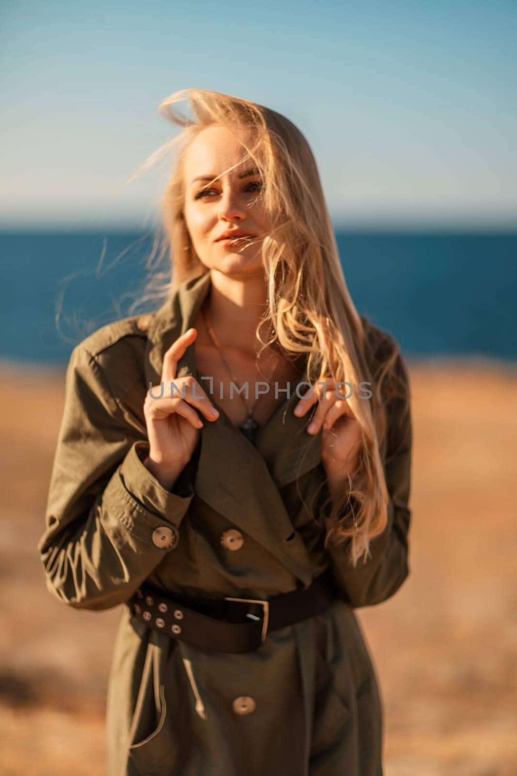 Portrait blonde sea cape. Calm young blonde in a khaki raincoat stands on the seashore by Matiunina