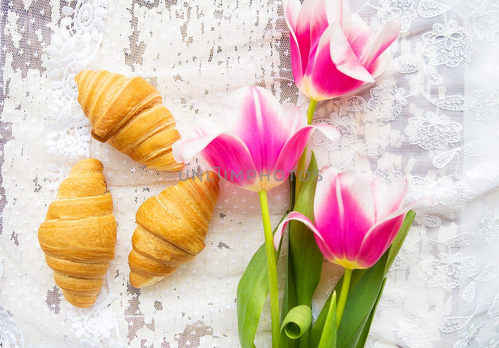 Three croissants and bright pink tulips on lace tablecloth.