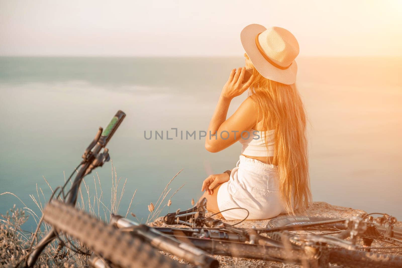 Happy female cyclist gazing at the stunning view of mountains and sea while sitting on a rock and chatting on the phone during an adventurous bike ride