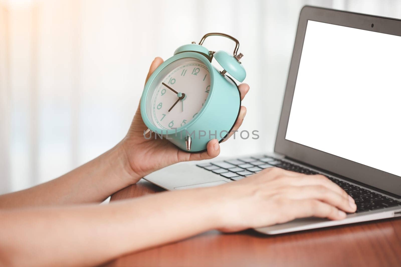 Asian woman's hand holding alarm clock to check the time on desk with a computer laptop for the concept of work, study and time management.