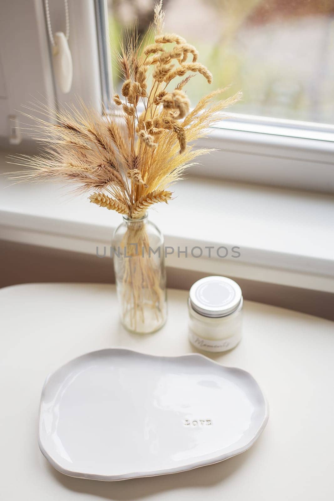 A white plate made of handmade clay, a candle, a vase stands near the window