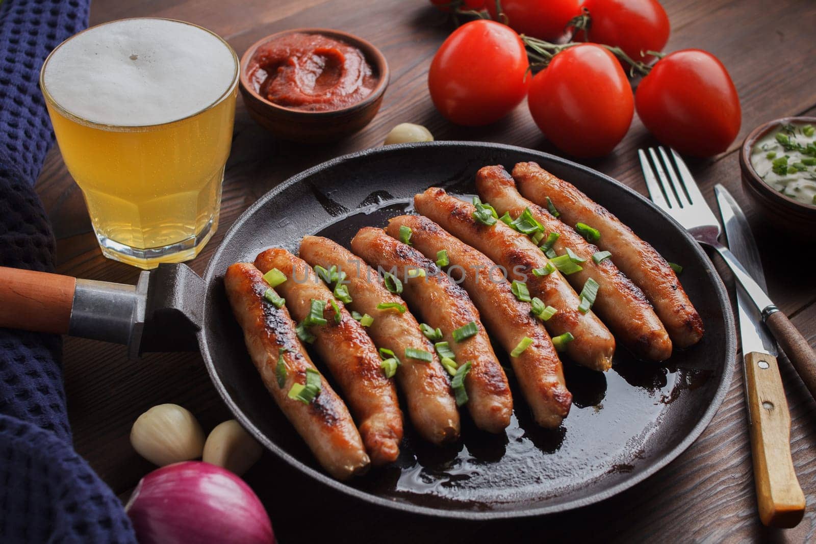 Delicious sausages cooked in a frying pan with vegetables, various sauces and a glass of beer on a wooden table.