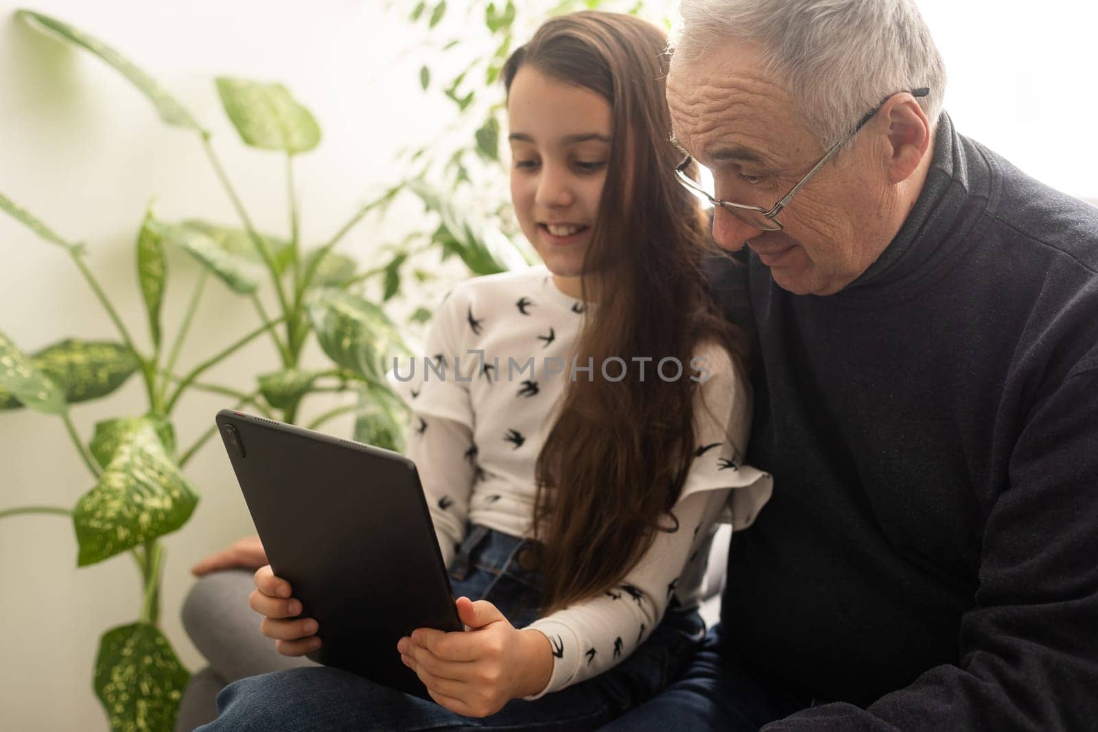 Cute little girl and her handsome grandpa are smiling while sitting on couch at home. Girl is using a tablet