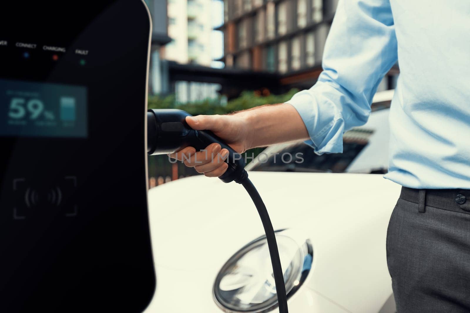 Closeup progressive man holding EV charger plug from public charging station for electric vehicle with background of residential building as concept eco-friendly sustainability energy car concept.