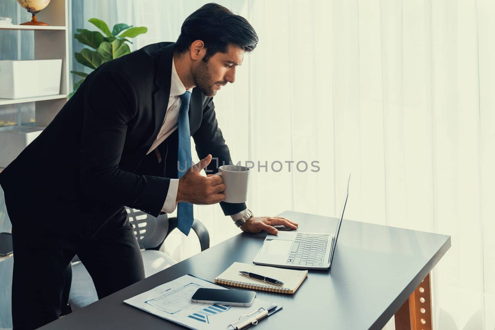 Modern professional businessman at modern office desk using laptop to work with coffee in his hand. Diligent office worker working on computer notebook in his office work space. fervent