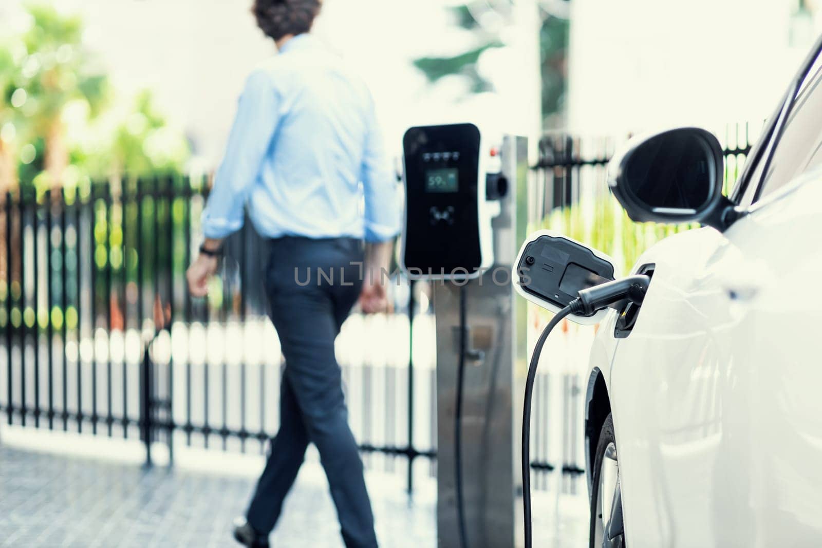 Progressive eco-friendly concept of focus parking EV car at public electric-powered charging station in city center with blur businessman walking in the background.