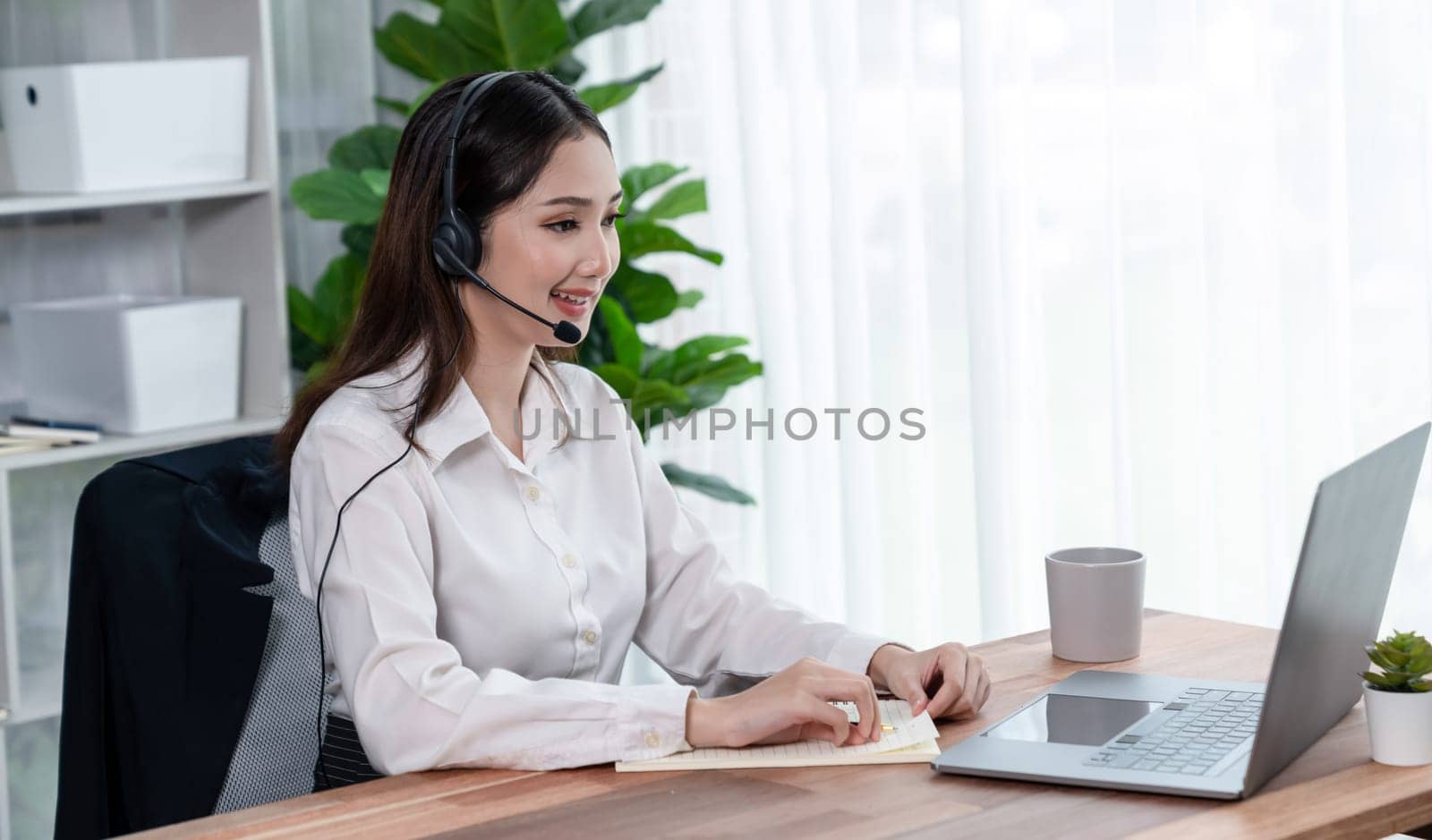 Enthusiastic asian call center with headset and microphone working on her laptop by biancoblue