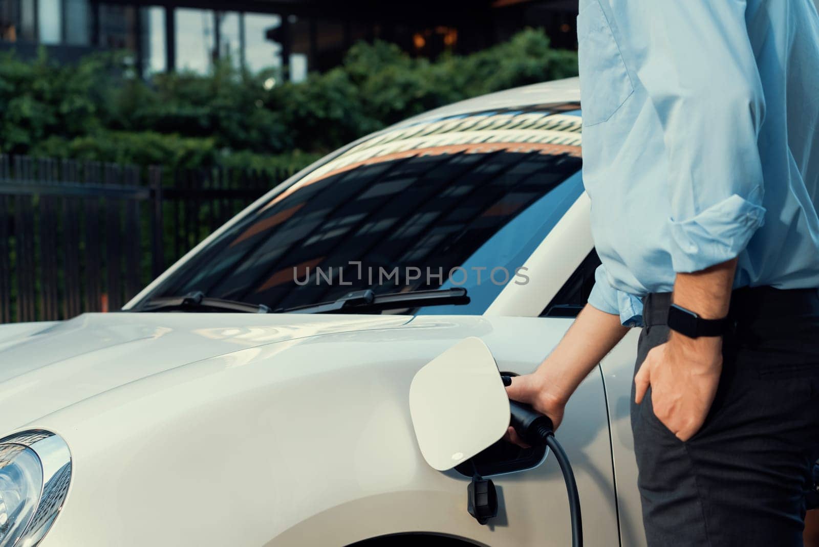 Closeup progressive man holding EV charger plug from public charging station for electric vehicle with background of residential building as concept eco-friendly sustainability energy car concept.