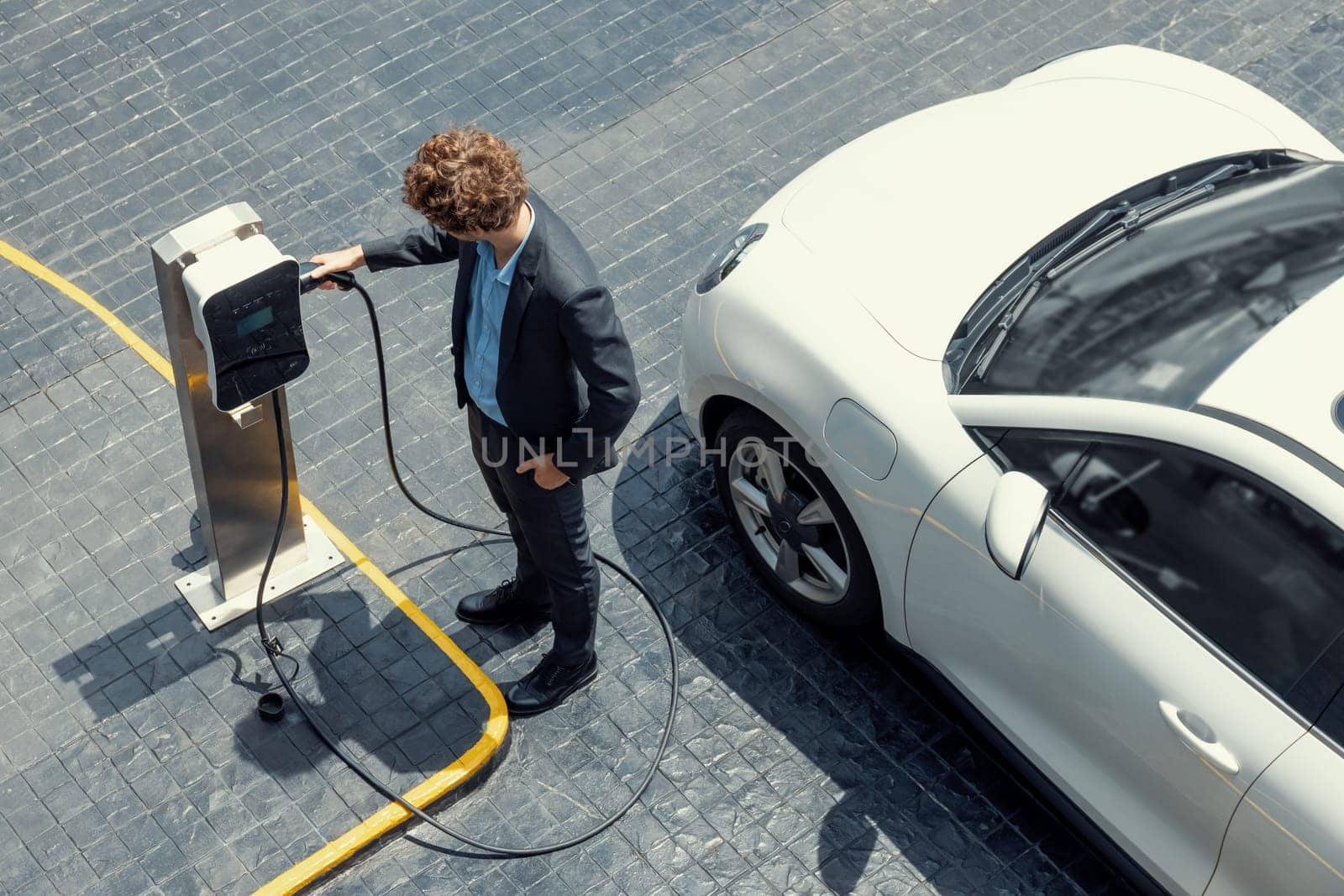 Aerial view of progressive businessman in black formal suit with his electric vehicle recharging battery at public car park charging station as vehicle powered by sustainable energy concept.