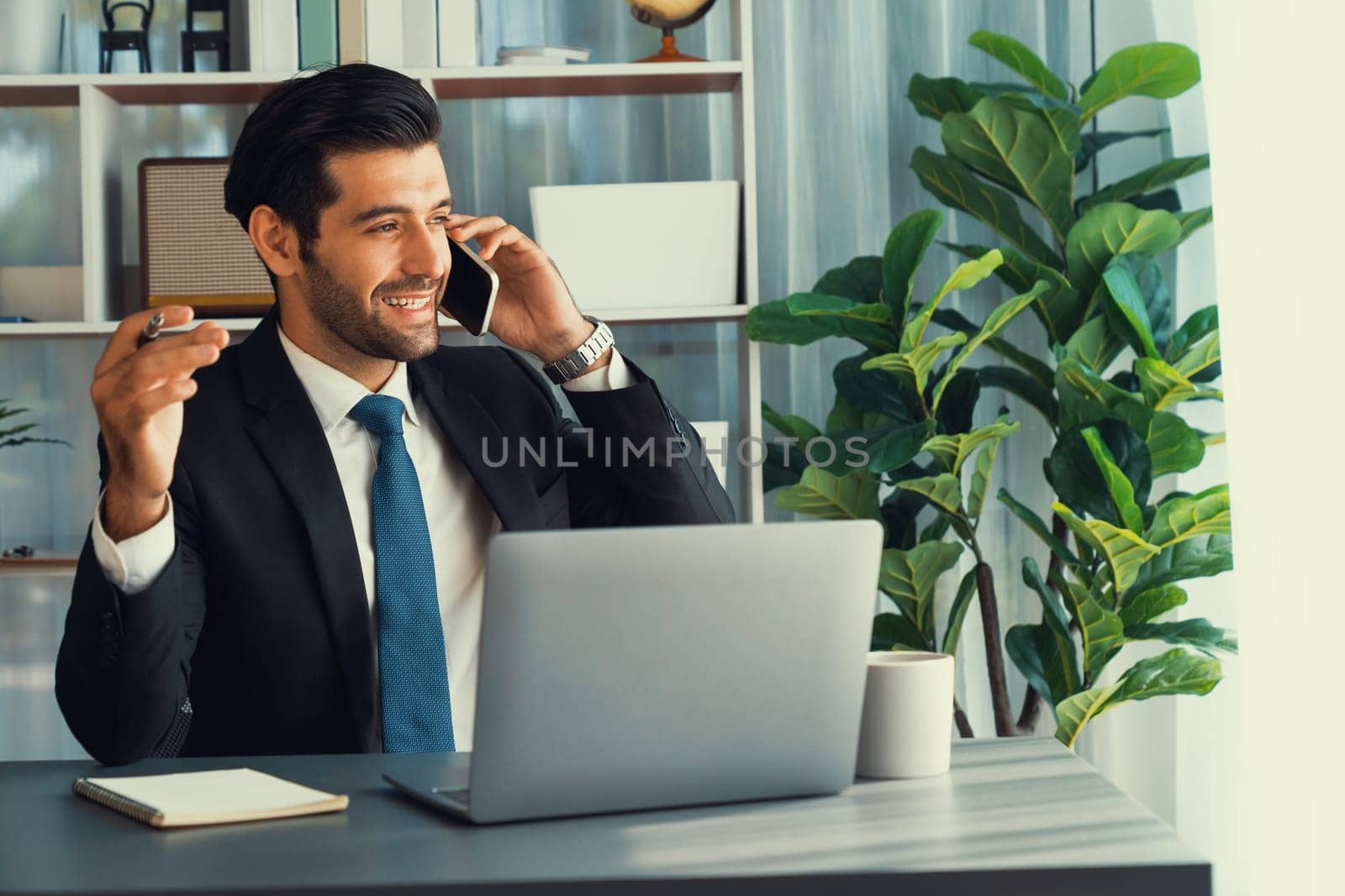 Diligent businessman busy talking on the phone call with clients while working with laptop in his office as concept of modern hardworking office worker lifestyle with mobile phone. Fervent