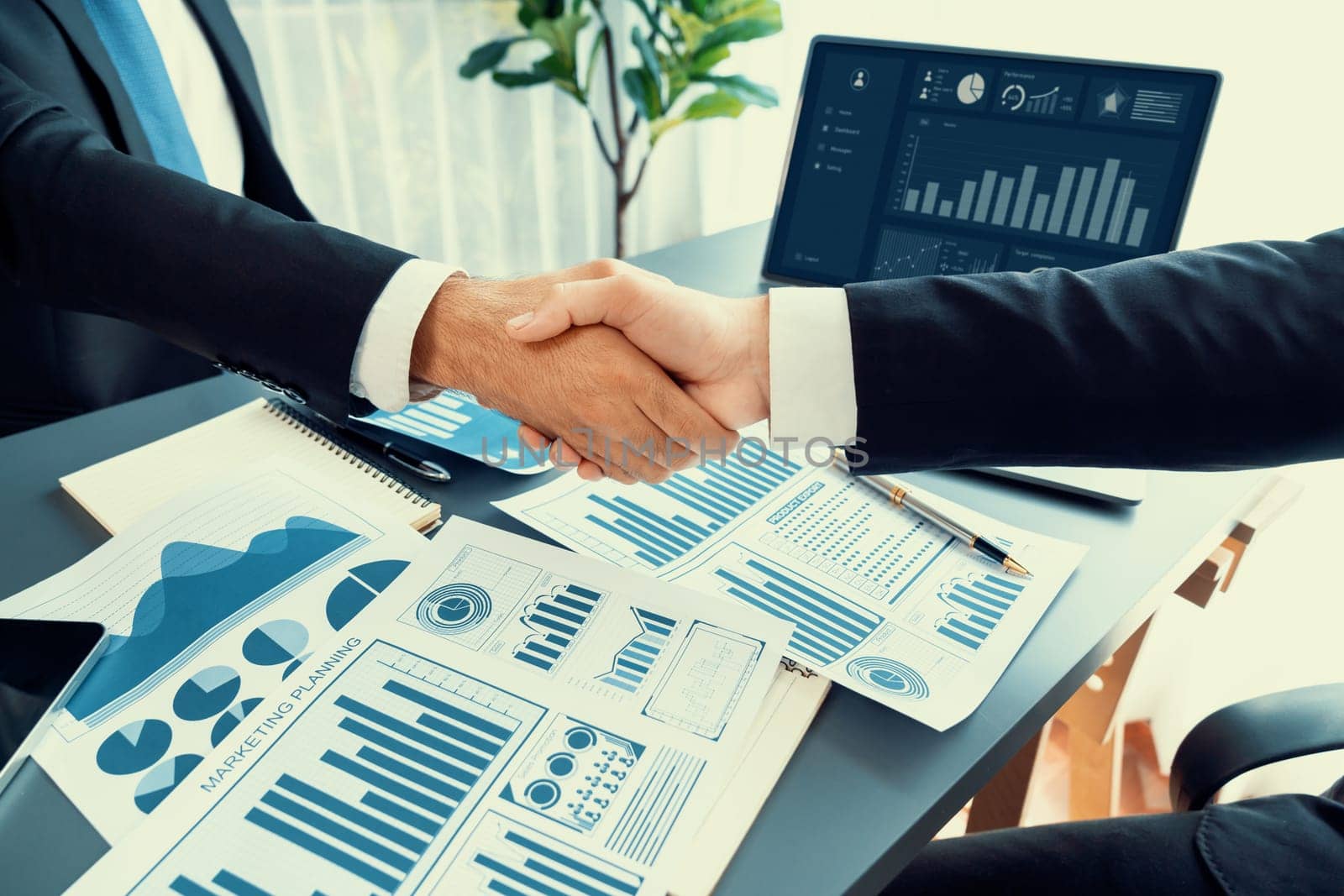 Closeup professional businessman shaking hands over desk in modern office after successfully analyzing pile of dashboard data paper as teamwork and integrity handshake in workplace concept. fervent