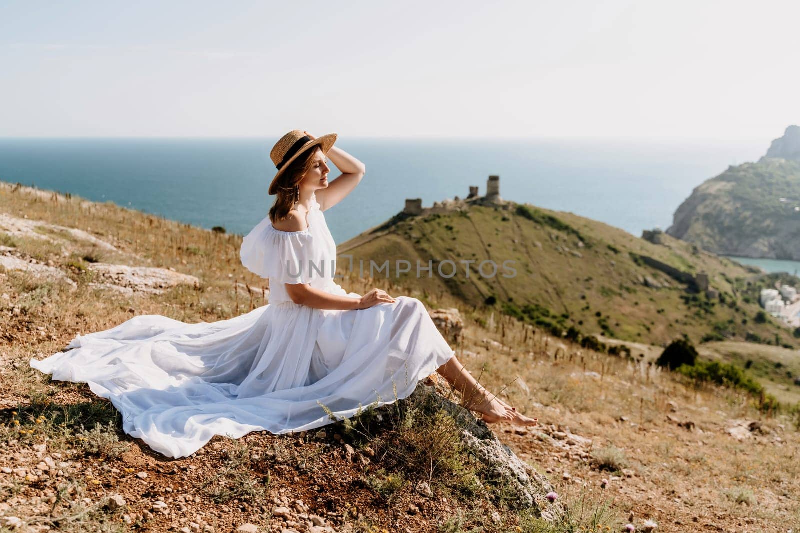 Happy woman in a white dress and hat stands on a rocky cliff above the sea, with the beautiful silhouette of hills in thick fog in the background. by Matiunina