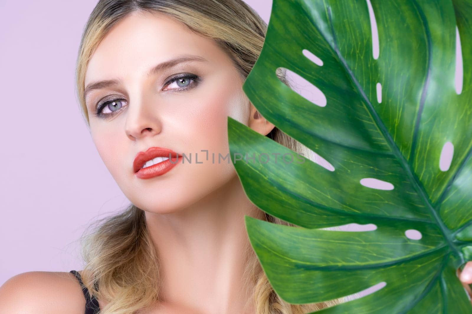Closeup woman with perfect clean skin and alluring flawless natural soft facial makeup holding green leave monstera. Natural skincare treatment beauty or spa concept in pink isolated background.