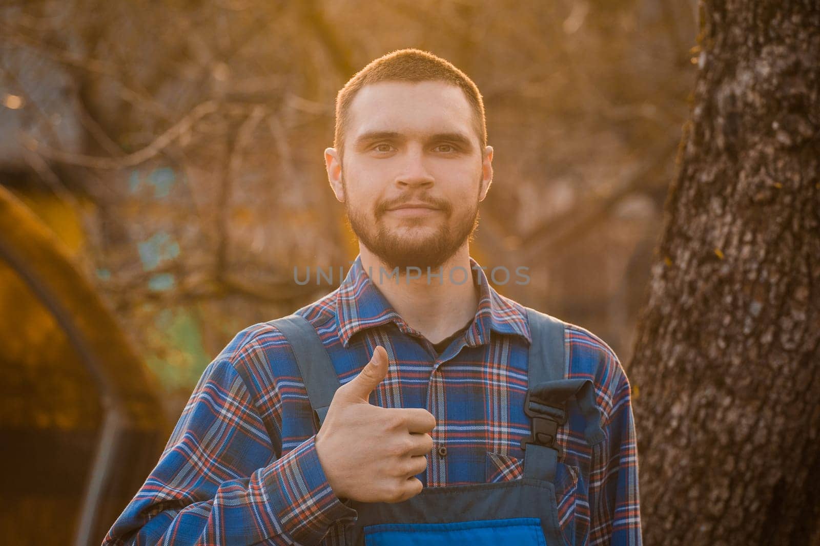 Farmer satisfied european appearance rural portrait at sunset with beard, shirt and overalls looking at camera, showing cool gesture with hand finger outdoors by AYDO8