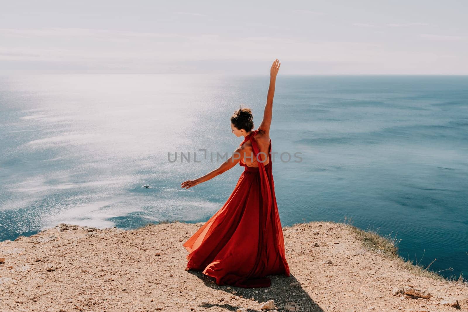 Woman in red dress on sea. Side view a Young beautiful sensual woman in a red long dress posing on a rock high above the sea on sunset. Girl on the nature on blue sky background. Fashion photo. by panophotograph