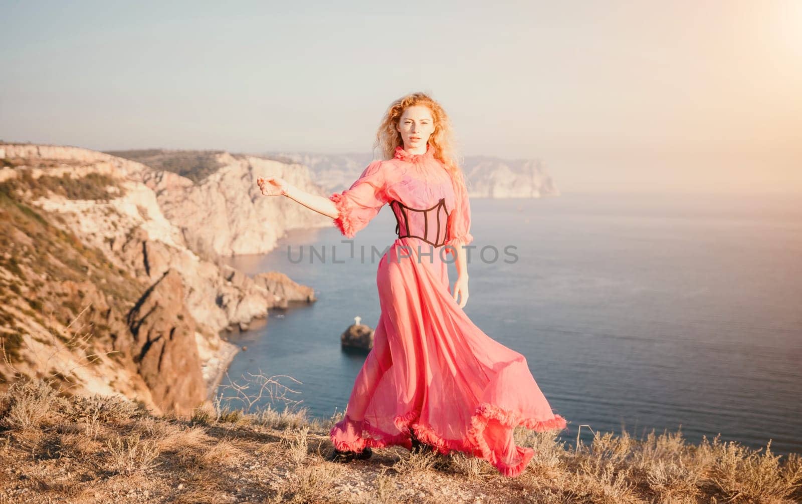 Curly redhead caucasian woman with freckles. Cute beautiful young blonde woman in a pink long dress posing on a volcanic rock high above the sea during sunset. Fashion travel conept by panophotograph