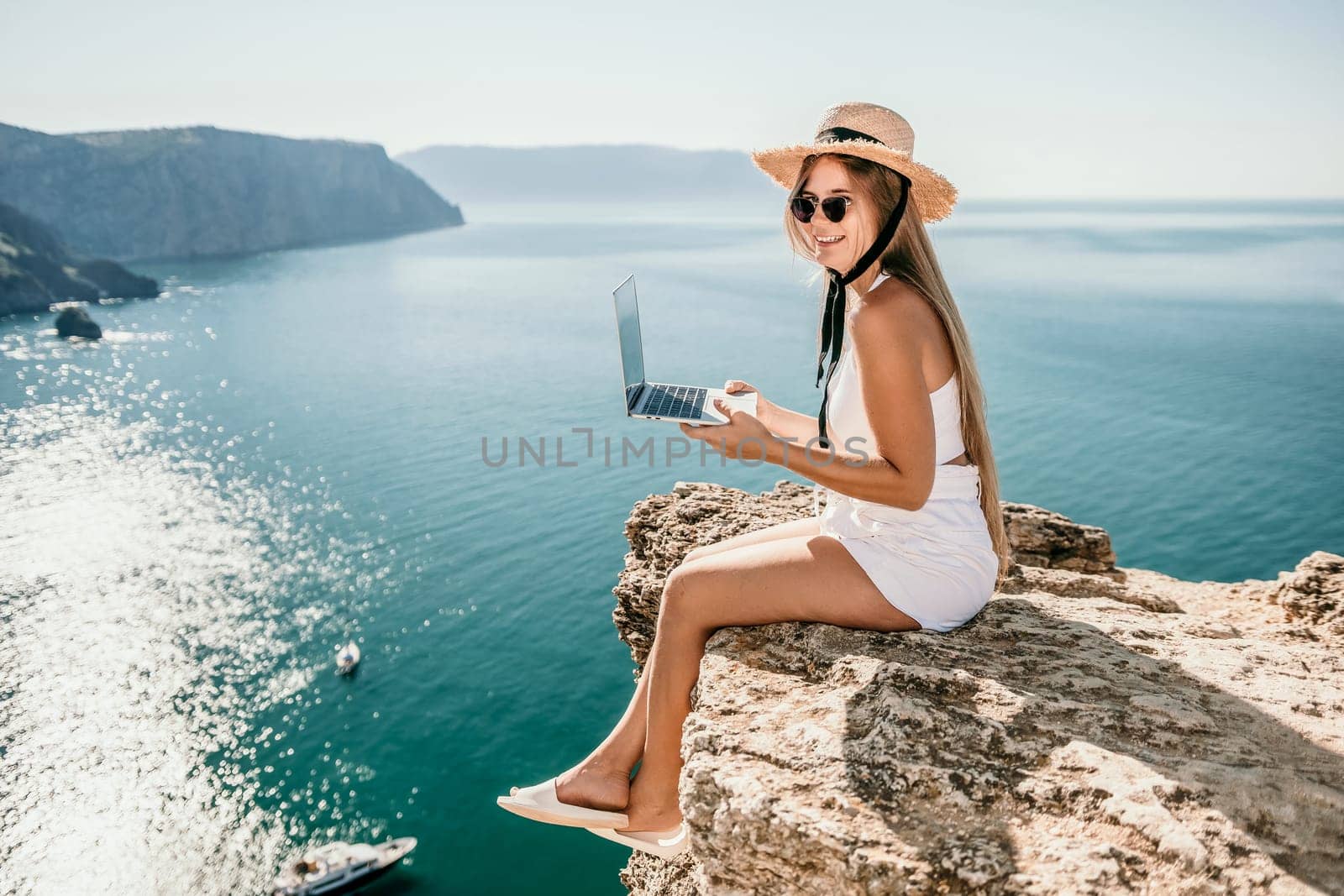 Woman laptop sea. Working remotely on seashore. Happy successful woman female freelancer in straw hat working on laptop by the sea at sunset. Freelance, remote work on vacation by panophotograph