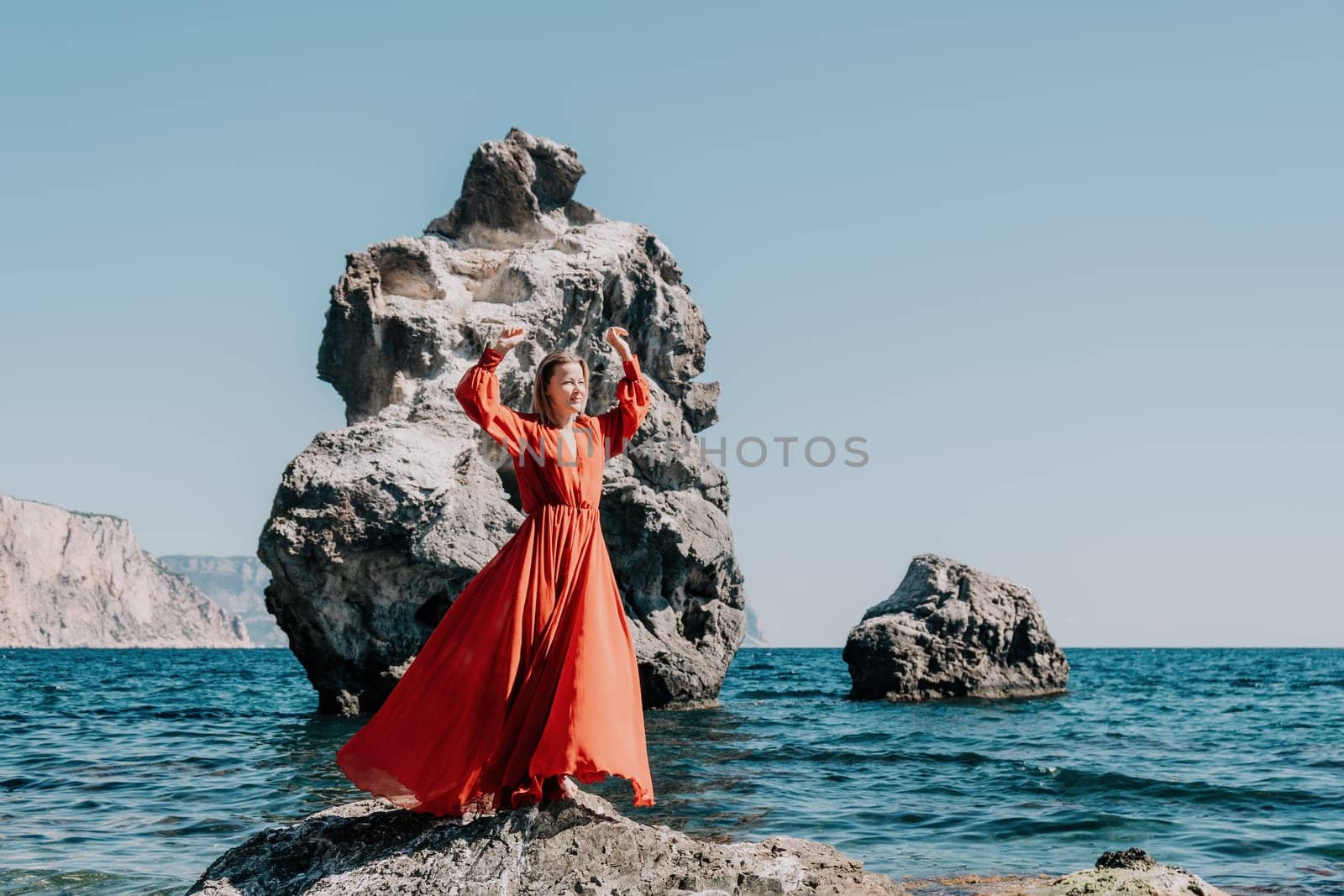 Woman travel sea. Young Happy woman in a long red dress posing on a beach near the sea on background of volcanic rocks, like in Iceland, sharing travel adventure journey