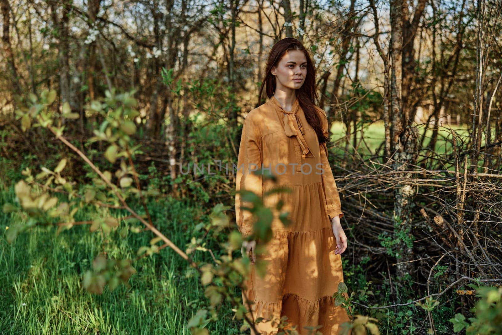 a woman with long hair walks in the shade near the trees, dressed in a long orange dress, enjoying the weather and the weekend, inspecting the trees. The theme of privacy with nature. High quality photo