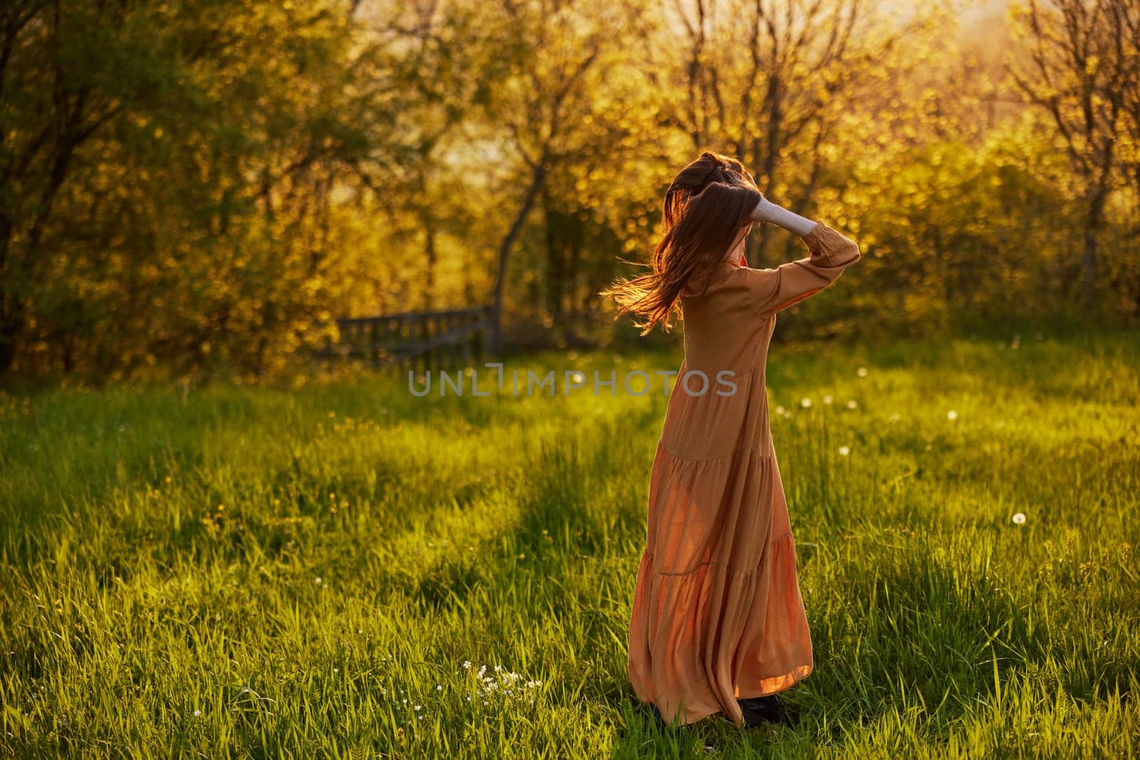 a slender woman with long hair stands in a field with her back to the camera, illuminated by the rays of the setting sun and happily poses enjoying the warm weather and rest by Vichizh