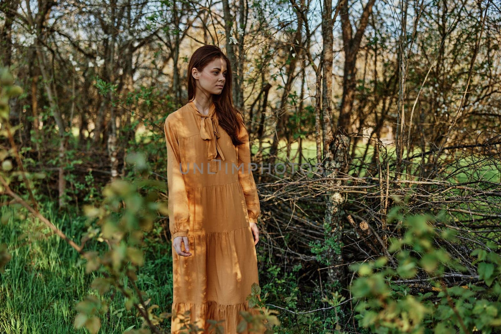 woman with long hair walks in the shade near the trees, dressed in a long orange dress, enjoying the weather and the weekend. The theme of privacy with nature, horizontal photography on the street by Vichizh