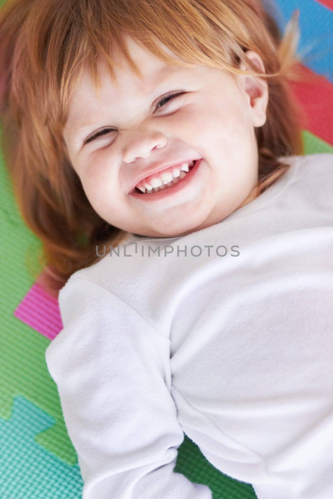 Young girl, laughing and happiness portrait of a baby on a home playpen ground with a smile. Ginger infant, kid laugh and happy in a house with joy, youth and positivity from childhood looking up by YuriArcurs