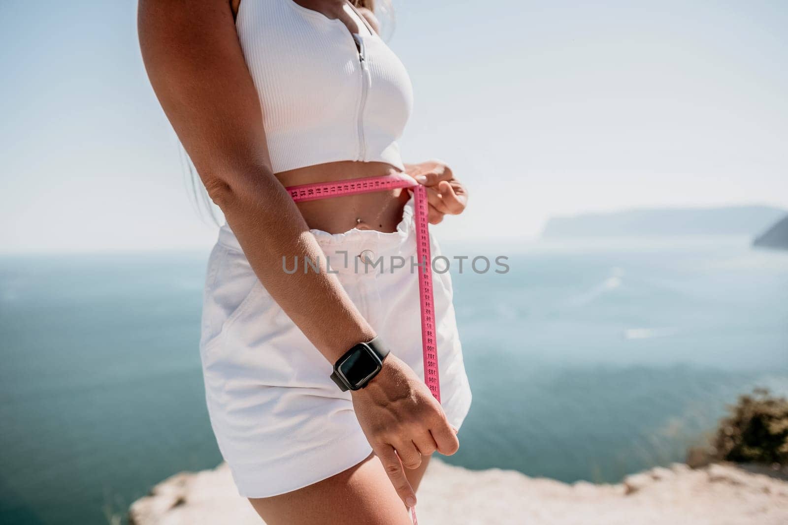 woman in white shorts holds in her hands pink measuring tape on her waist near the azure sea. Concept of detox and diet by panophotograph