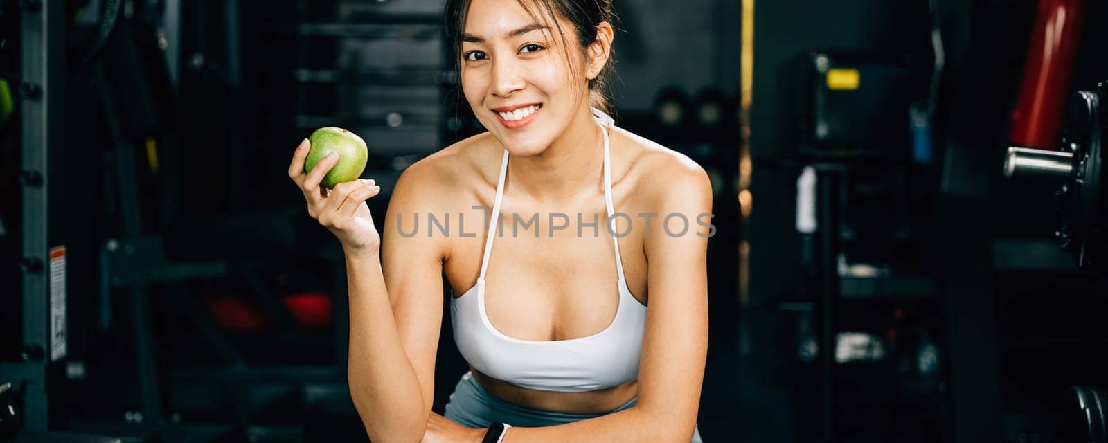 A female runner holds a green apple in a gym, reminding viewers of the importance of healthy food choices for achieving fitness goals. Healthy fitness and eating lifestyle concept