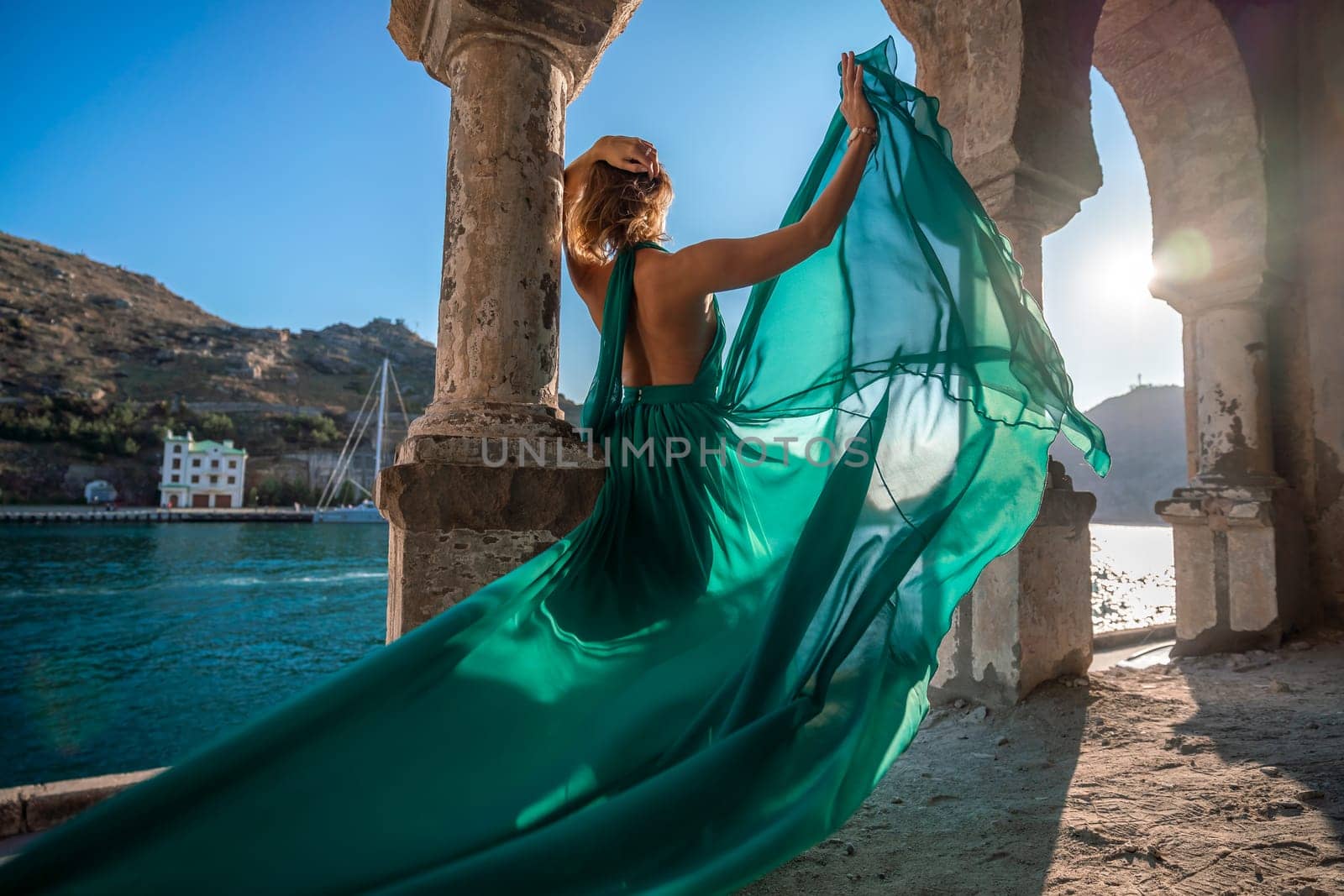 Rear view of a happy blonde woman in a long mint dress posing against the backdrop of the sea in an old building with columns. Girl in nature against the blue sky