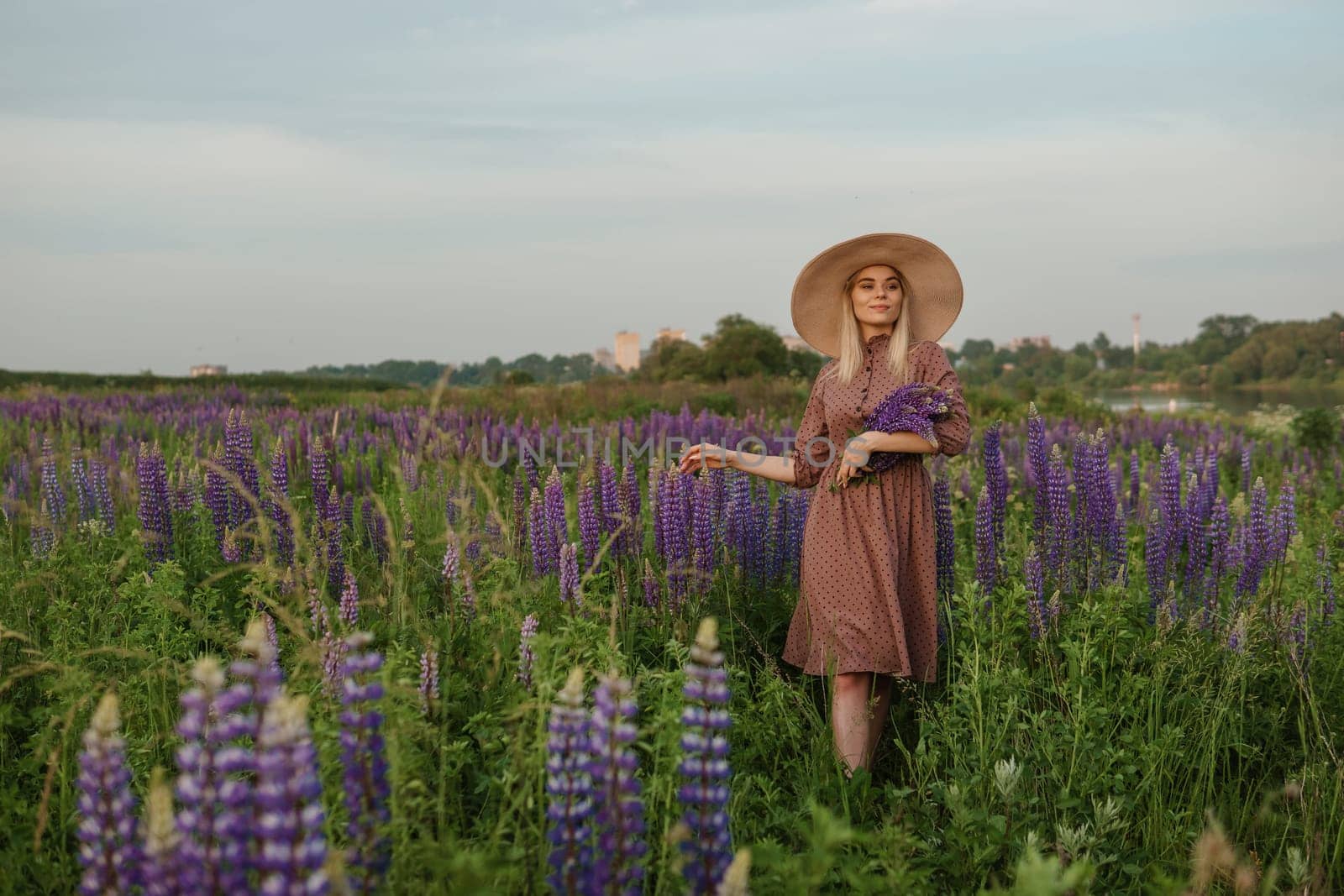 A beautiful woman in a straw hat walks in a field with purple flowers. A walk in nature in the lupin field.