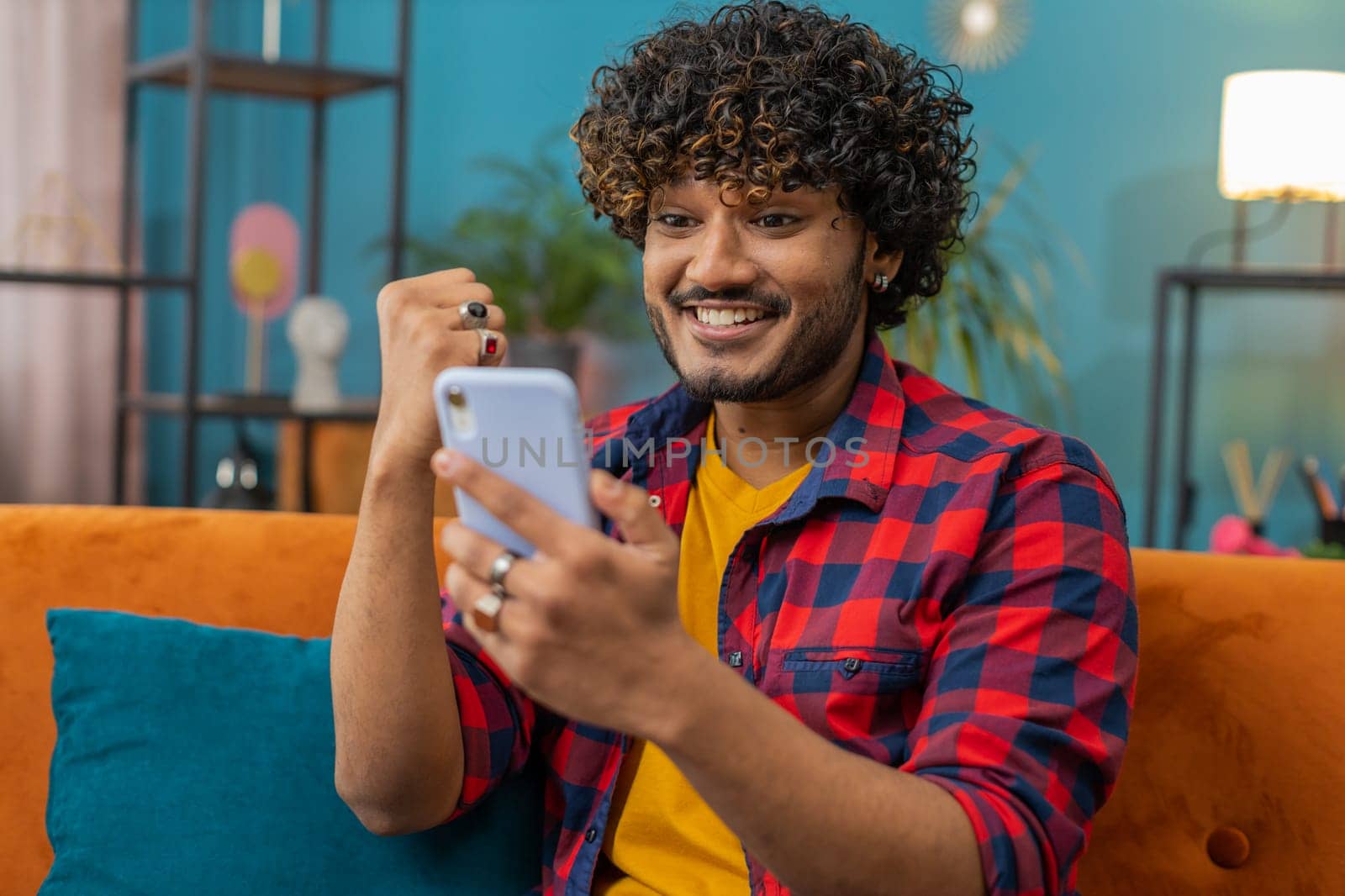 Happy indian man use mobile smartphone typing browsing say Wow yes found out great big win good news celebrate lottery jackpot doing winner gesture. Hindu young guy at home in room sitting on sofa