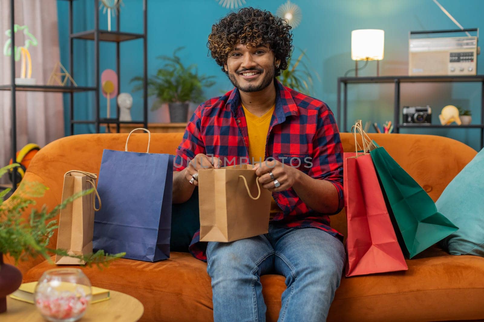 Young indian man happy shopaholic consumer came back home after shopping sale with bags. Portrait of hindu guy satisfied received parcels purchase from online order at home apartment room on sofa