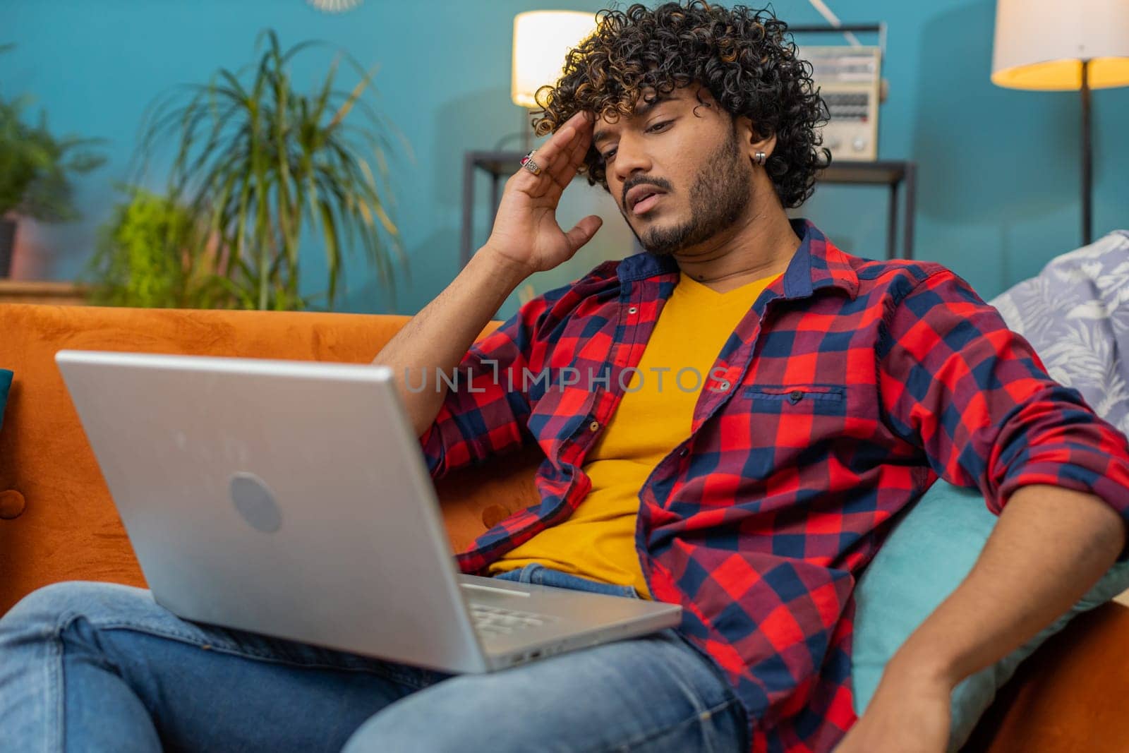 Tired freelancer indian man use laptop, suffering from headache problem tension and migraine, stress at home room. Hindu guy works on notebook, sends messages, makes online purchases, watching movies