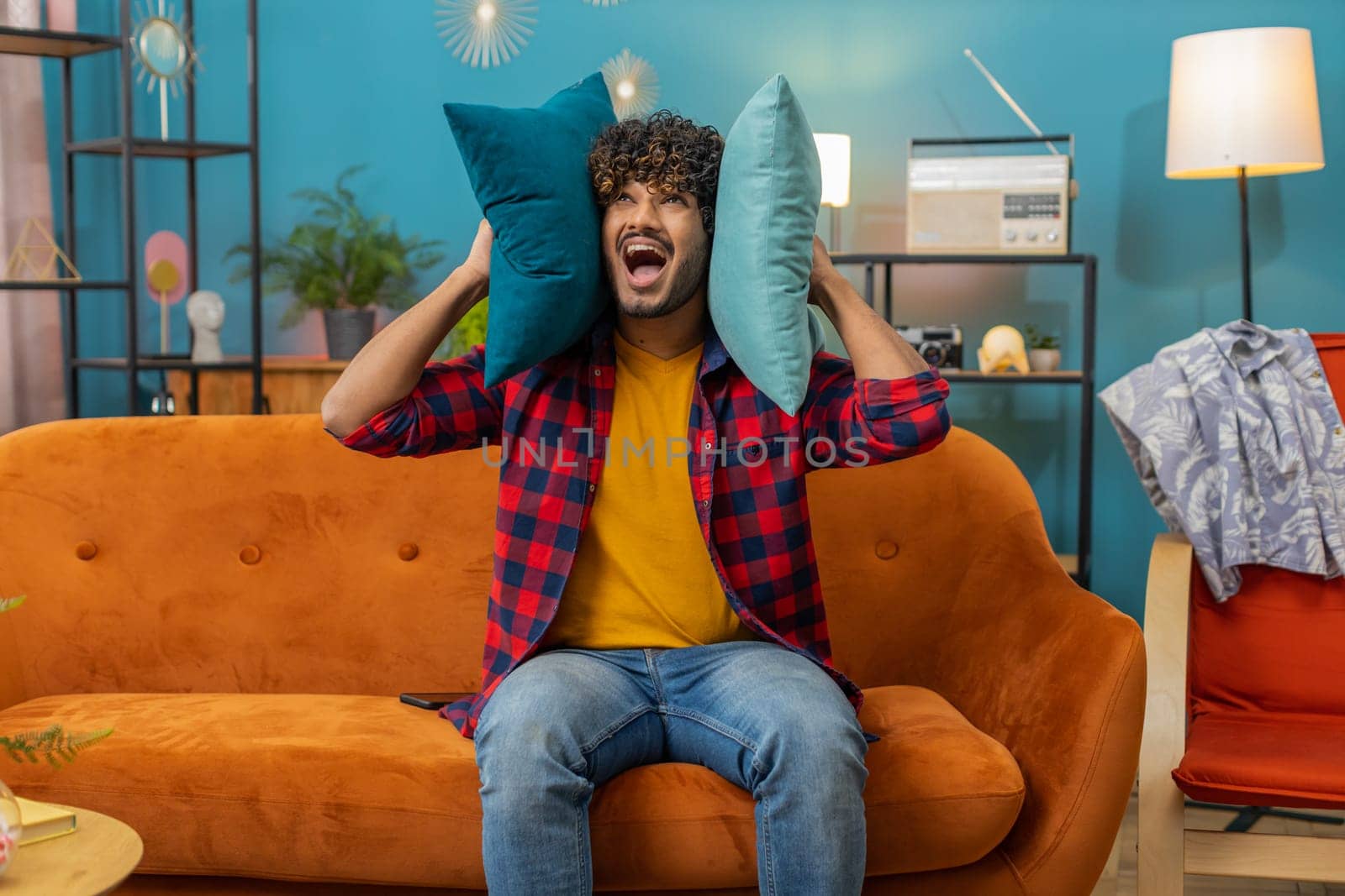 Repair work at neighbours. Irritated indian man relaxing on couch cover ears with pillows annoyed by noisy neighbors suffer from headache wish silence. Thin walls at home flat without sound insulation