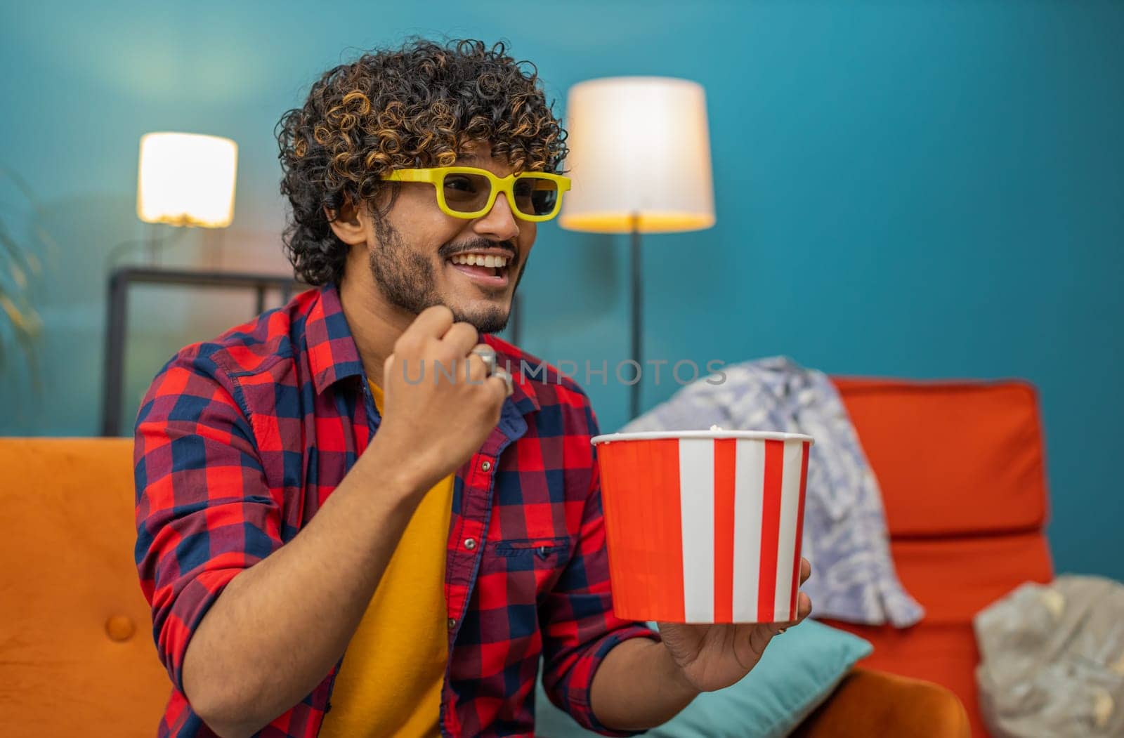 Excited indian man in 3D glasses sits on sofa eating popcorn snacks and watching interesting TV serial sport game, film, online social media movie content at home apartment. Guy enjoying entertainment