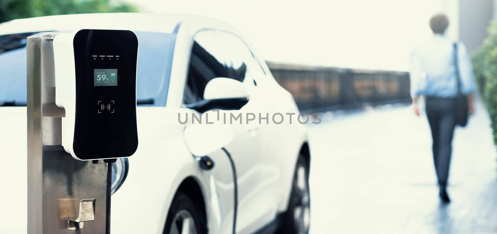 Progressive eco-friendly concept of focus parking EV car at public electric-powered charging station in city center with blur businessman walking in the background.