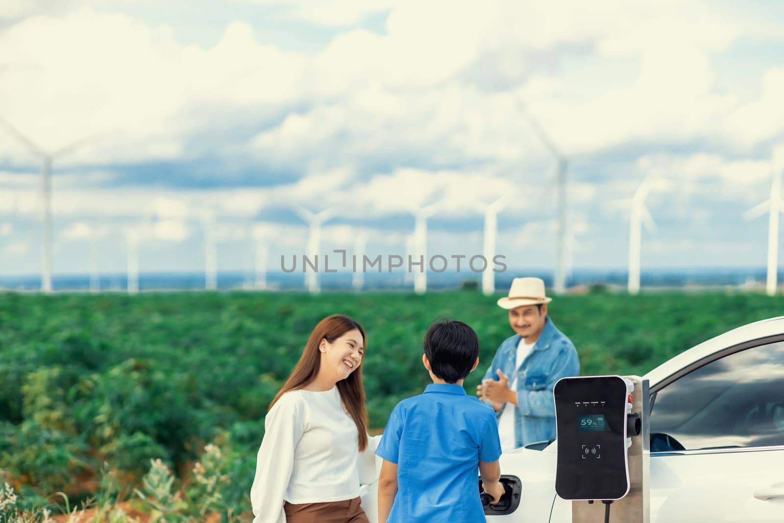 Concept of progressive happy family enjoying their time at wind farm with electric vehicle. Electric vehicle driven by clean renewable energy from wind turbine generator for charging station.